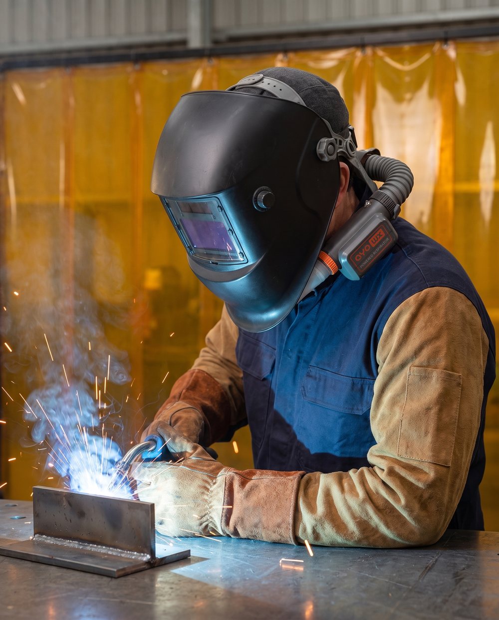 Welder MIG welding under a lowered welding helmet while wearing an AYO WX around-neck powered air-purifying respirator